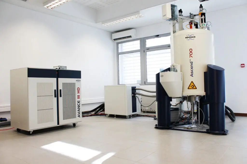 Advanced NMR spectrometer setup in a bright laboratory, featuring a large cylindrical magnet and control console.