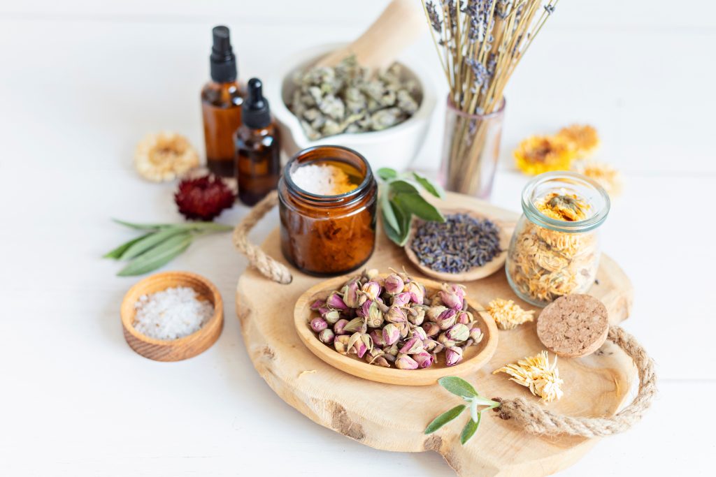 Assorted herbs and flowers in jars, with dried lavender, rosebuds, and essential oil bottles on a wooden platter.