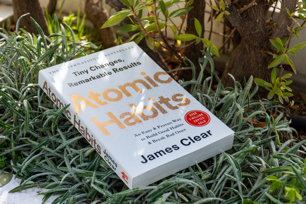 Atomic Habits" book by James Clear resting on green foliage, highlighting its title and subtitle in bold lettering.