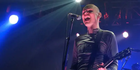 A bald musician passionately sings into a microphone, holding a guitar, with vibrant stage lights in the background.