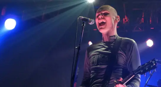A bald musician passionately sings into a microphone, holding a guitar, with vibrant stage lights in the background.