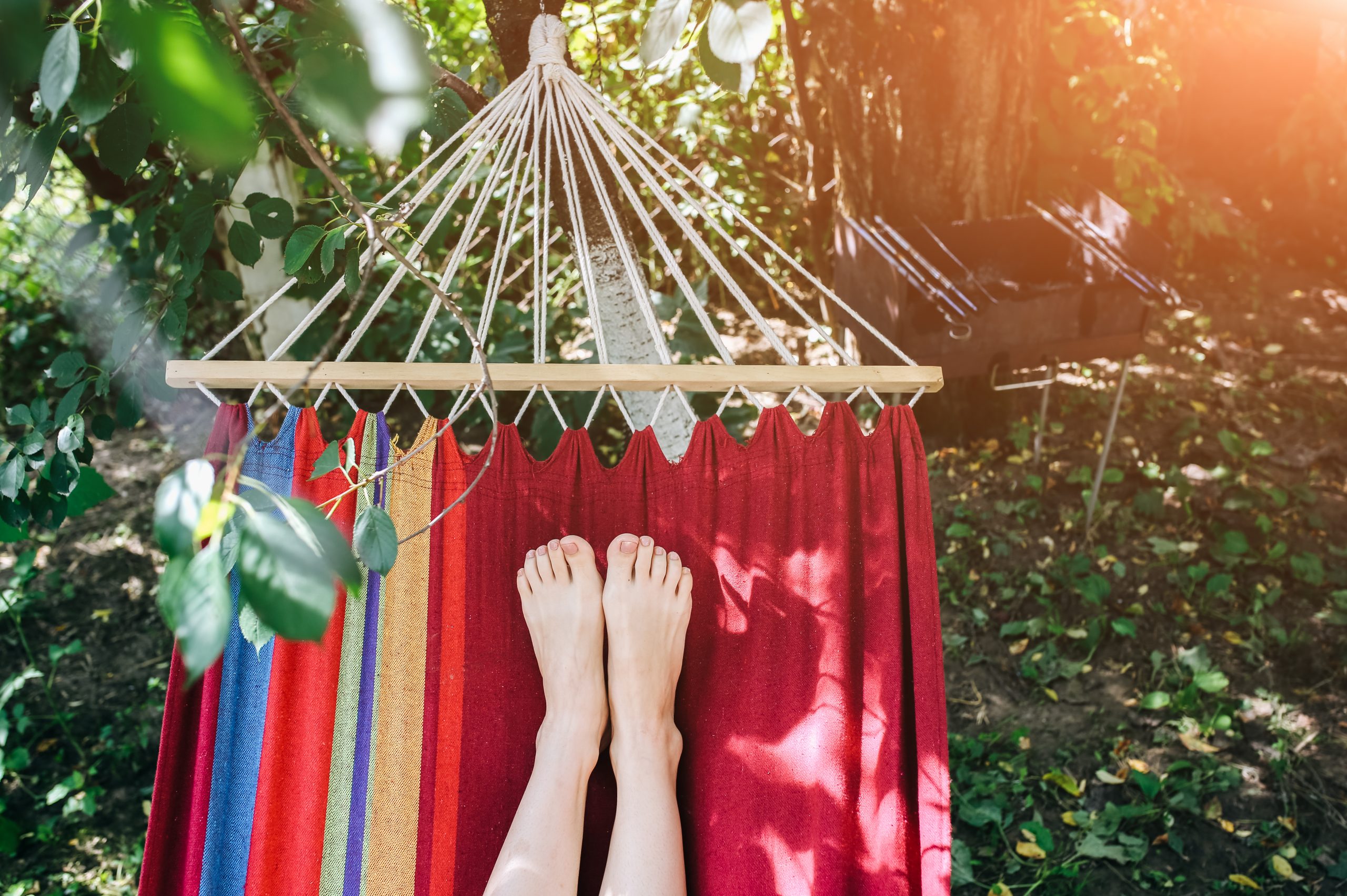Bare feet resting on a colorful hammock, surrounded by lush greenery and dappled sunlight. A grill is visible nearby.