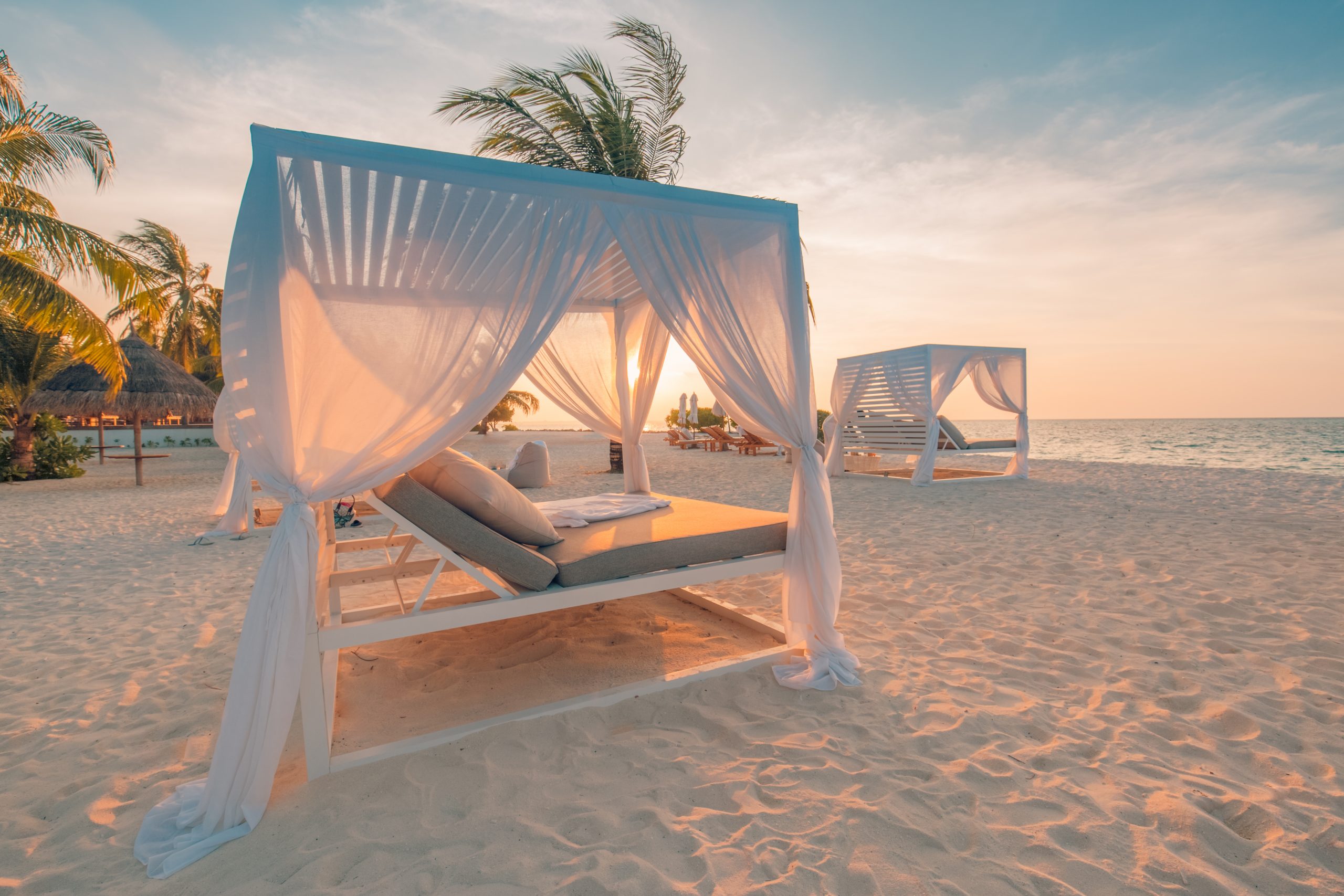 Beach cabanas with sheer curtains at sunset, surrounded by palm trees and soft sand, creating a serene tropical atmosphere.