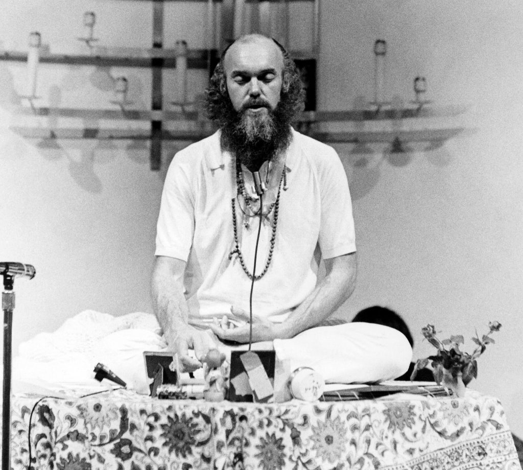 A bearded man in white clothing meditates on a patterned cloth, surrounded by candles and small objects.