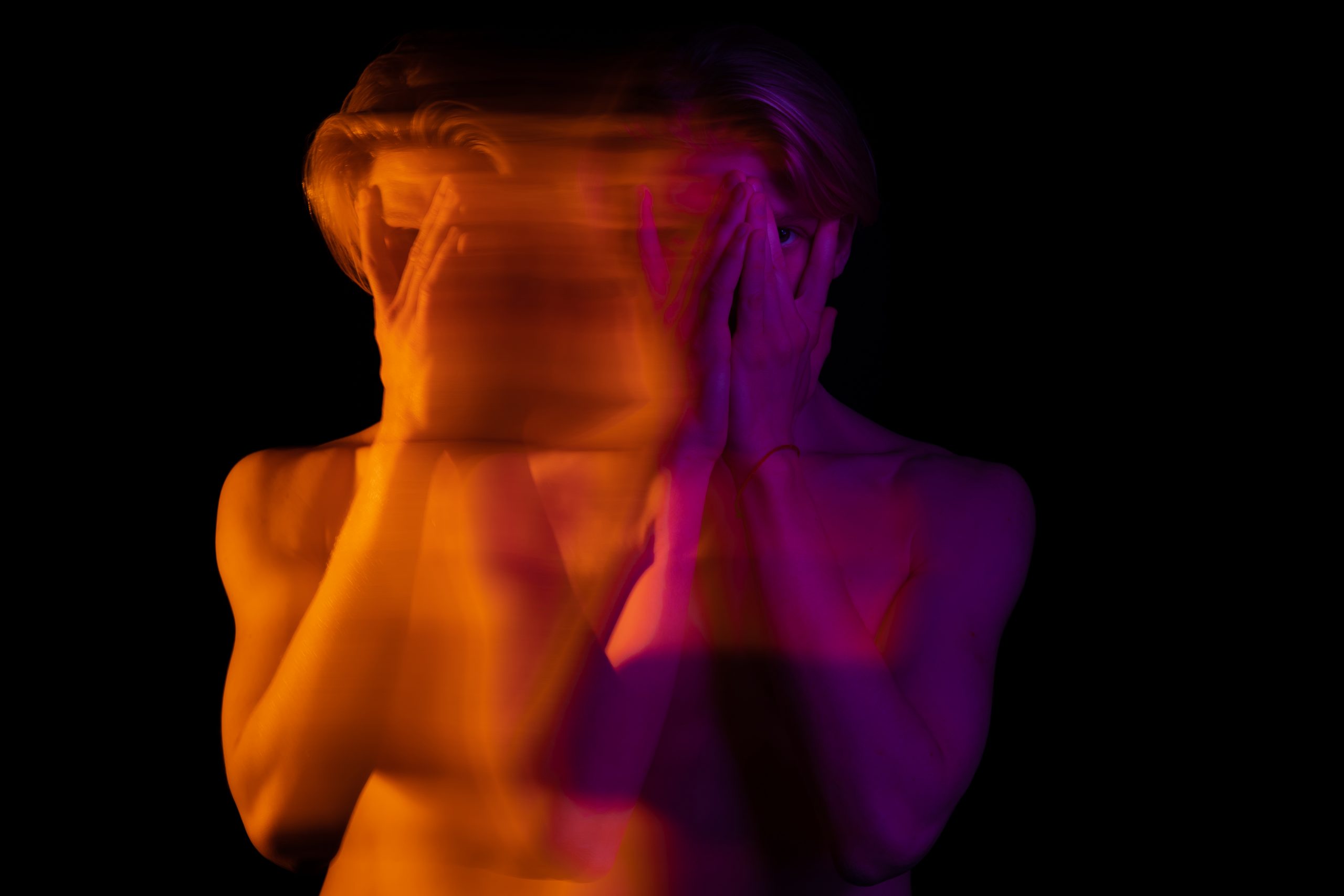 A blurred figure with hands covering their face, illuminated in vibrant orange and purple hues against a black background.