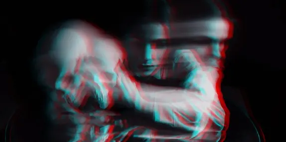 A blurred figure leans on a skull, creating a haunting, surreal atmosphere with contrasting shadows and colors.