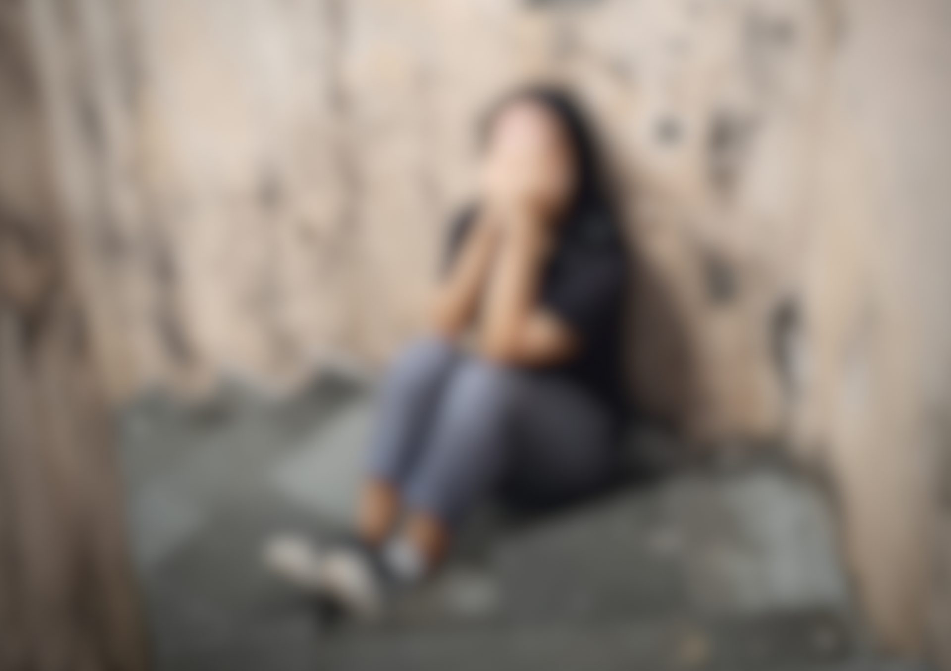 A blurred figure sits against a textured wall, hands covering their face, conveying a sense of distress or sadness.