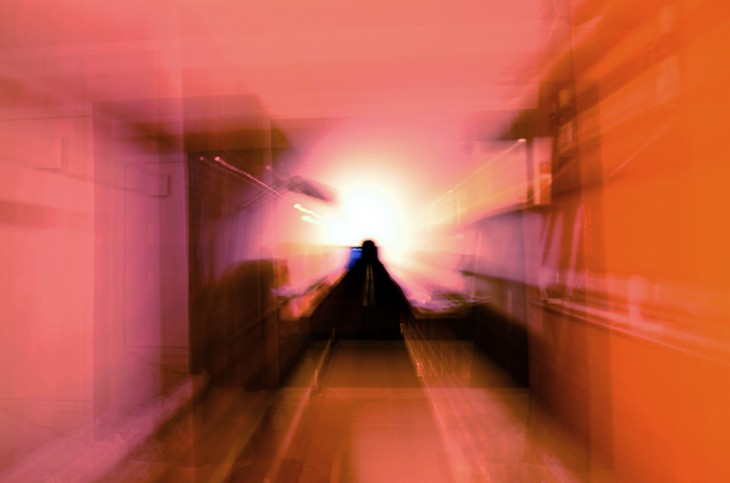 A blurred figure stands in a corridor, illuminated by a bright light, creating an abstract, dreamlike atmosphere.