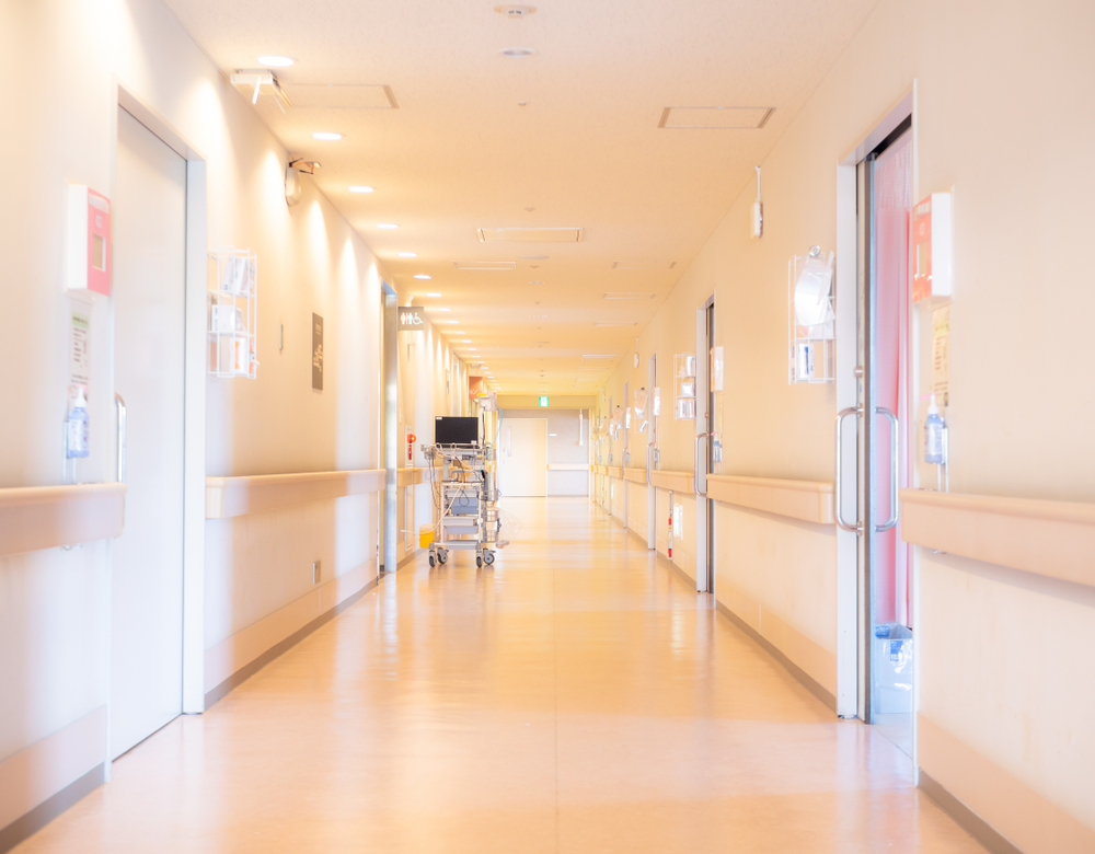 Bright hospital corridor with light-colored walls, medical equipment on wheels, and doors lining both sides.