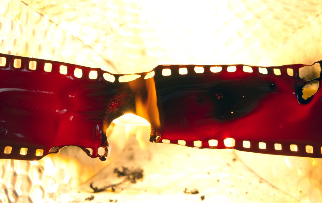 Burned film strip with charred edges and vibrant red color, illuminated against a bright background.