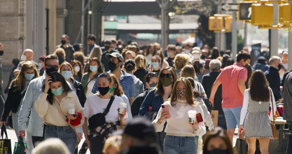 Busy city street filled with pedestrians wearing masks, holding coffee cups, and shopping bags on a sunny day.
