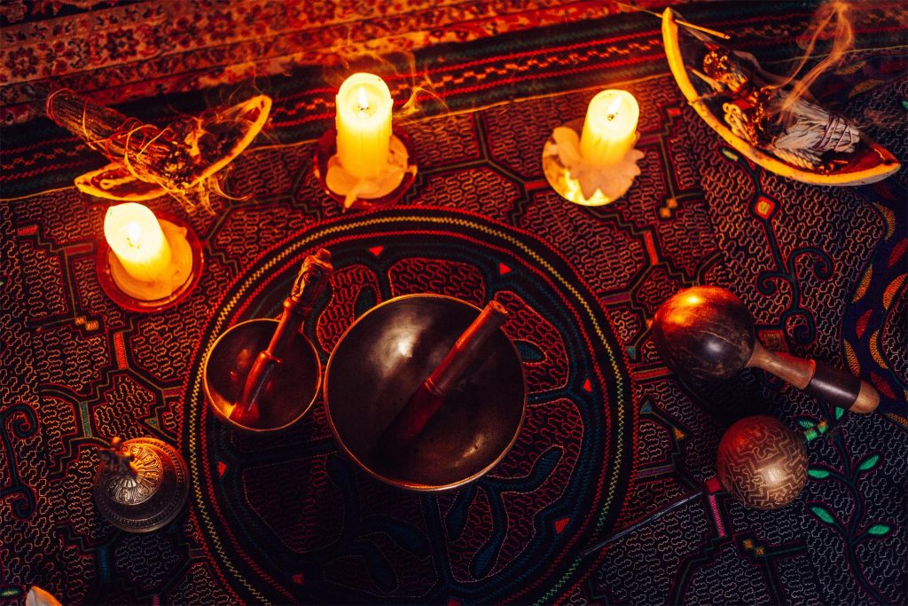 Candles glow softly around bowls and instruments on a richly patterned textile, creating a mystical ambiance.
