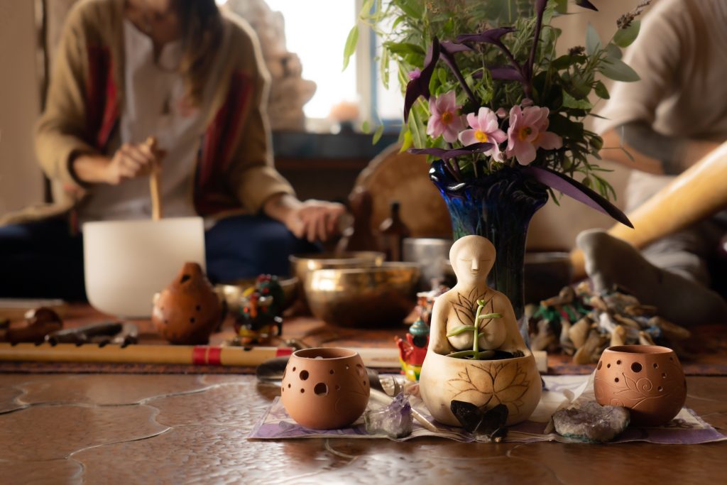 Ceramic figure surrounded by flowers, crystals, and musical instruments in a serene, artistic setting.