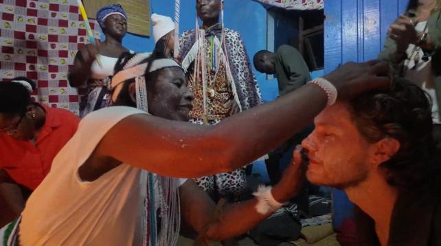 A ceremonial scene with participants in traditional attire, one person performing a ritual on another's face.