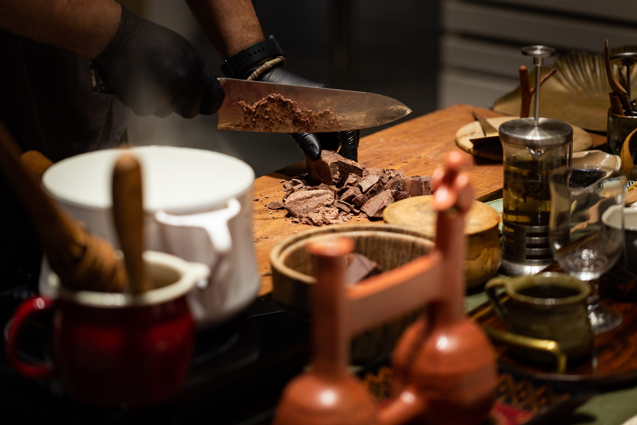 A chef in black gloves chops chocolate on a wooden board, surrounded by various kitchen tools and ingredients.
