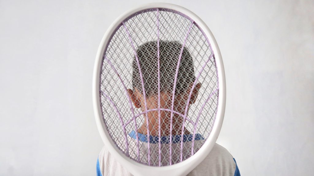 Child's head partially obscured by a large tennis racket, facing away against a plain background.