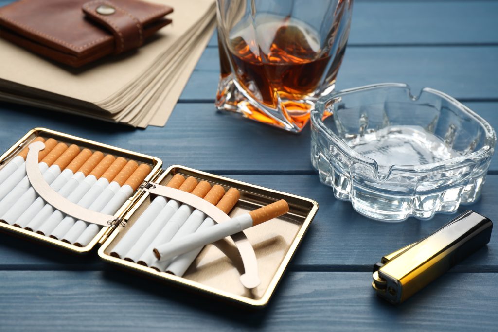 Cigarettes in a gold case, an empty ashtray, a glass of whiskey, and a brown wallet on a blue wooden table.