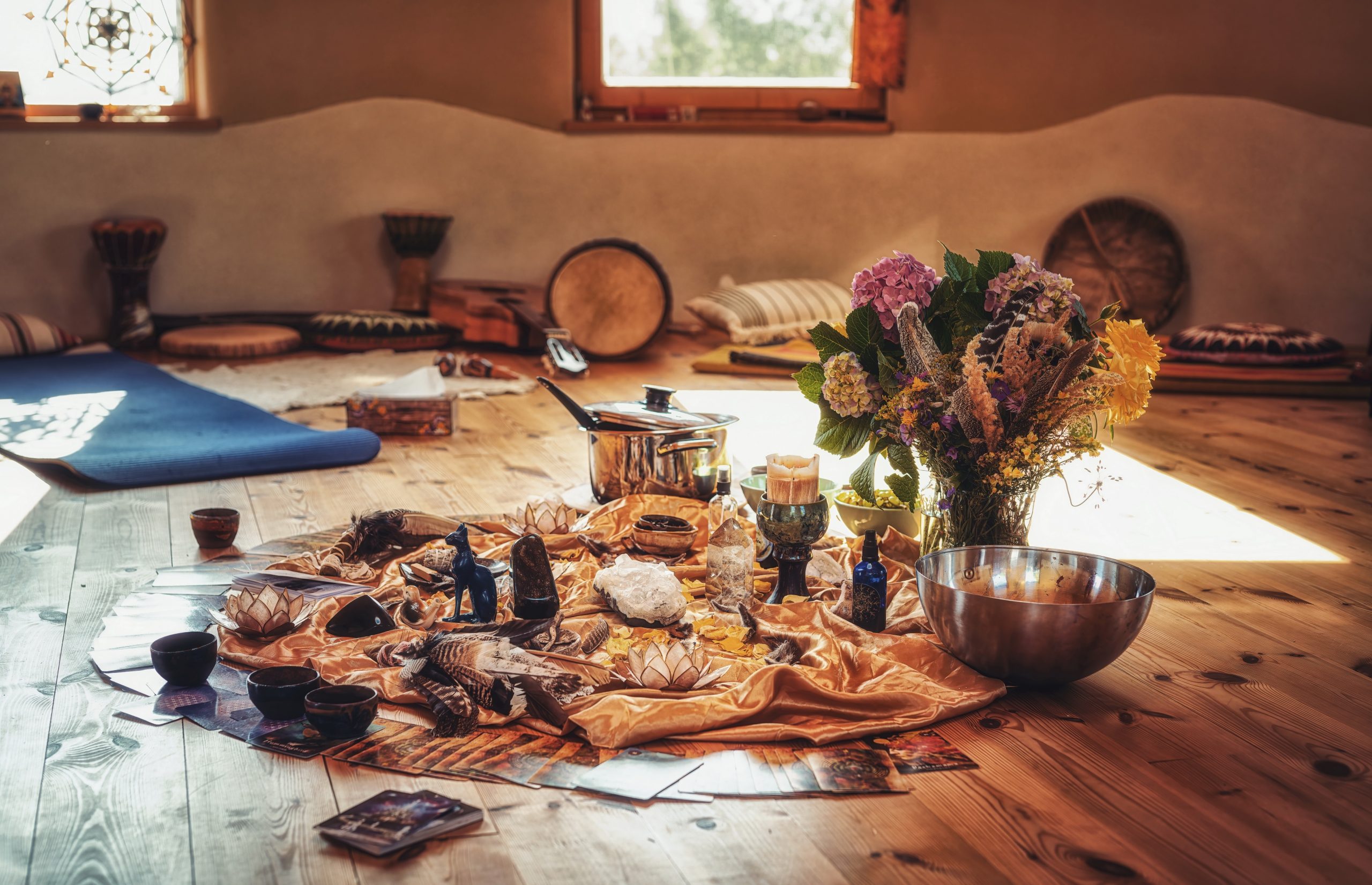 A circular arrangement of colorful fabrics, candles, flowers, and various artifacts on a wooden floor in a serene space.