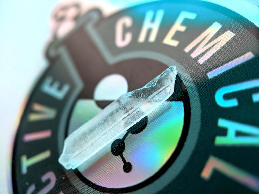 Clear crystal resting on a colorful chemical logo, reflecting light with a holographic sheen.