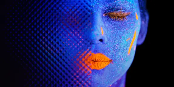 A close-up of a face painted in vibrant blue and orange hues, with glowing accents and a textured mesh overlay.