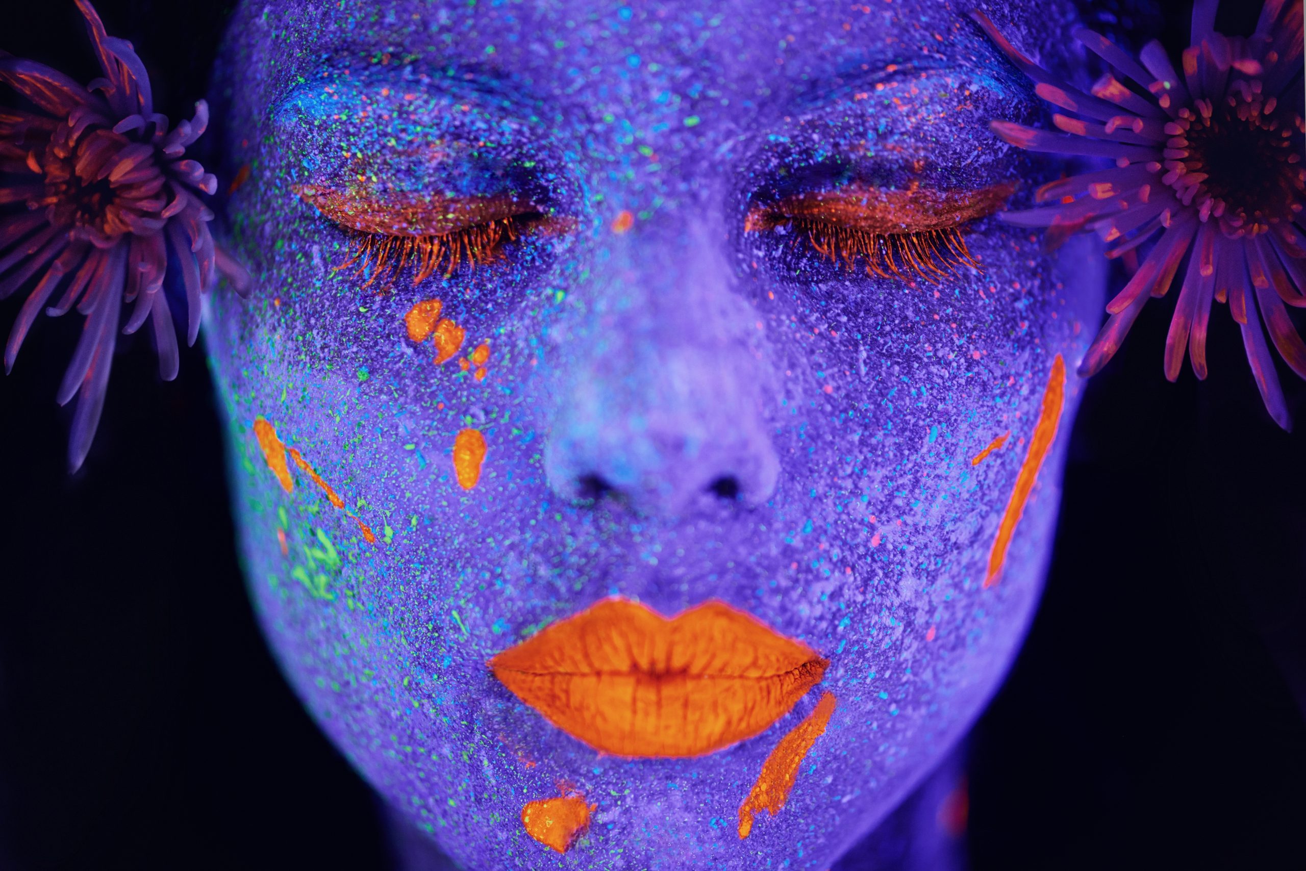 Close-up of a face with vibrant purple and orange body paint, adorned with flowers and glowing under UV light.