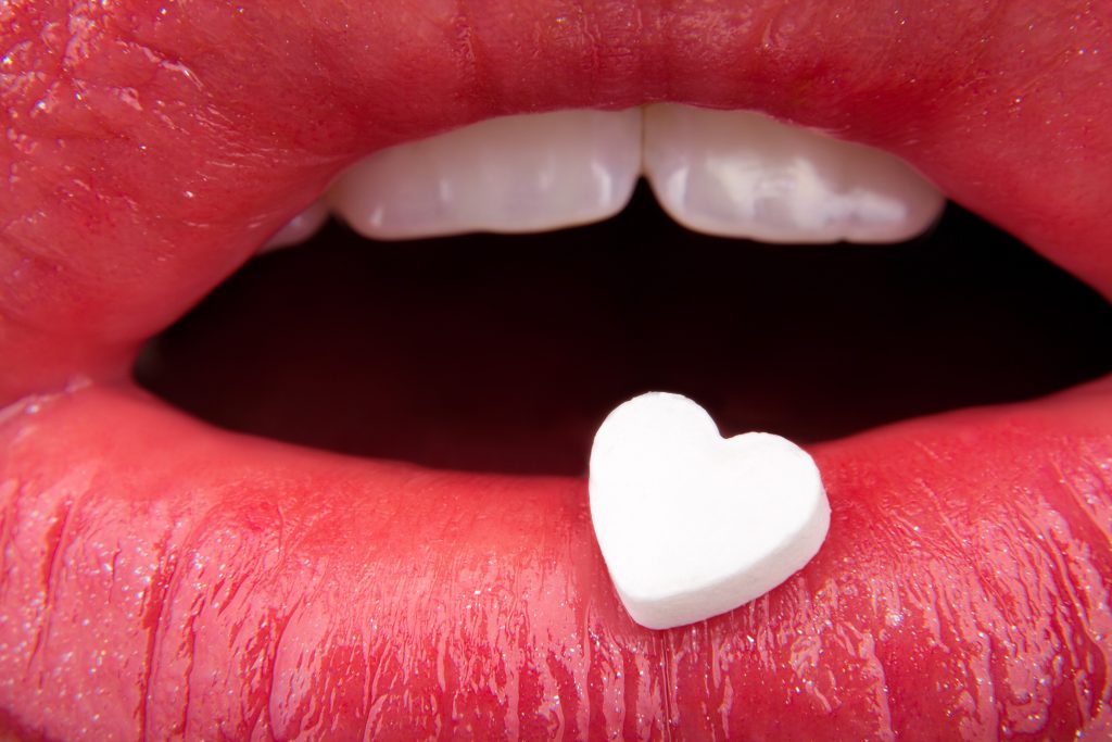 Close-up of glossy red lips holding a small white heart-shaped candy between them.