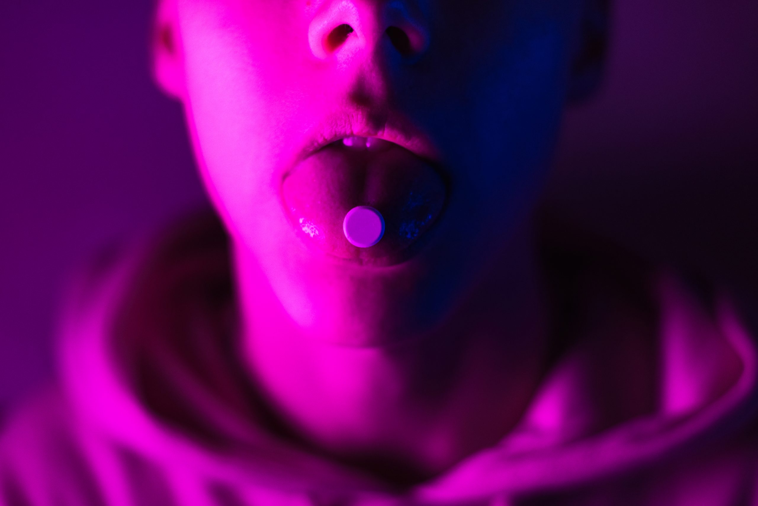 Close-up of a person with a pill on their tongue, illuminated by vibrant pink and purple lighting.