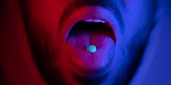 A close-up of a person's mouth with a blue pill resting on the tongue, illuminated by red and blue lighting.