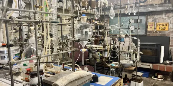 Cluttered laboratory filled with glassware, lab equipment, and shelves of chemicals, creating a busy scientific workspace.
