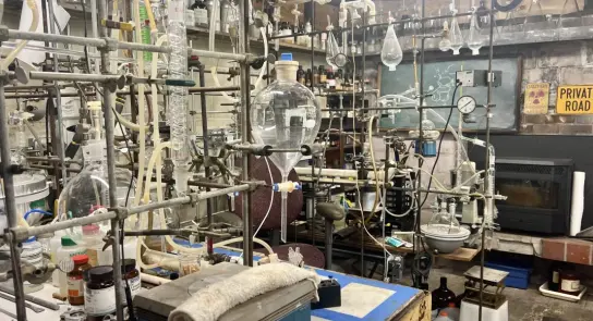 Cluttered laboratory filled with glassware, lab equipment, and shelves of chemicals, creating a busy scientific workspace.