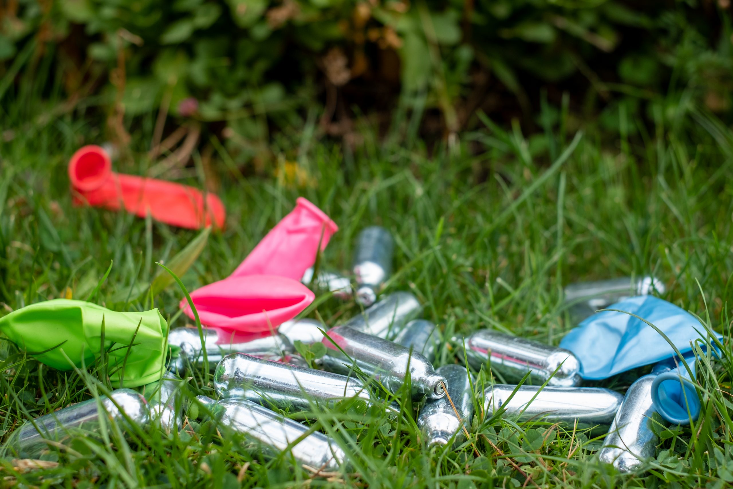 Colorful deflated balloons and silver whipped cream chargers scattered on green grass.