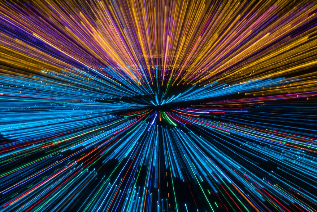 Colorful light streaks radiate outward in a dynamic burst, creating a sense of speed and energy against a dark background.