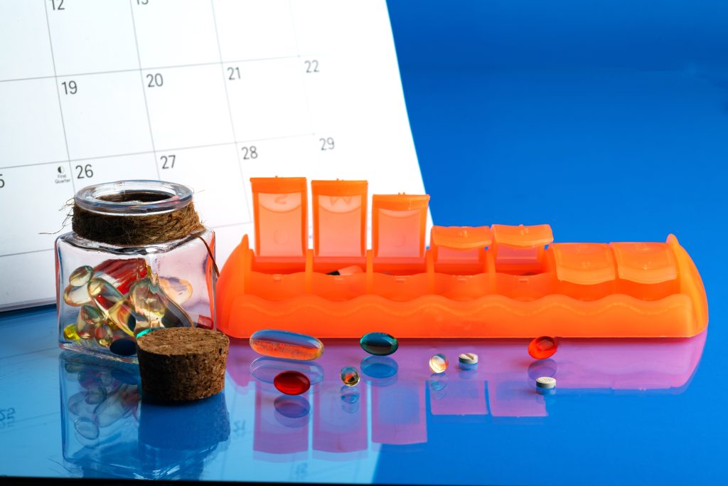 Colorful pills scattered around a clear jar and an orange pill organizer, with a calendar in the background.