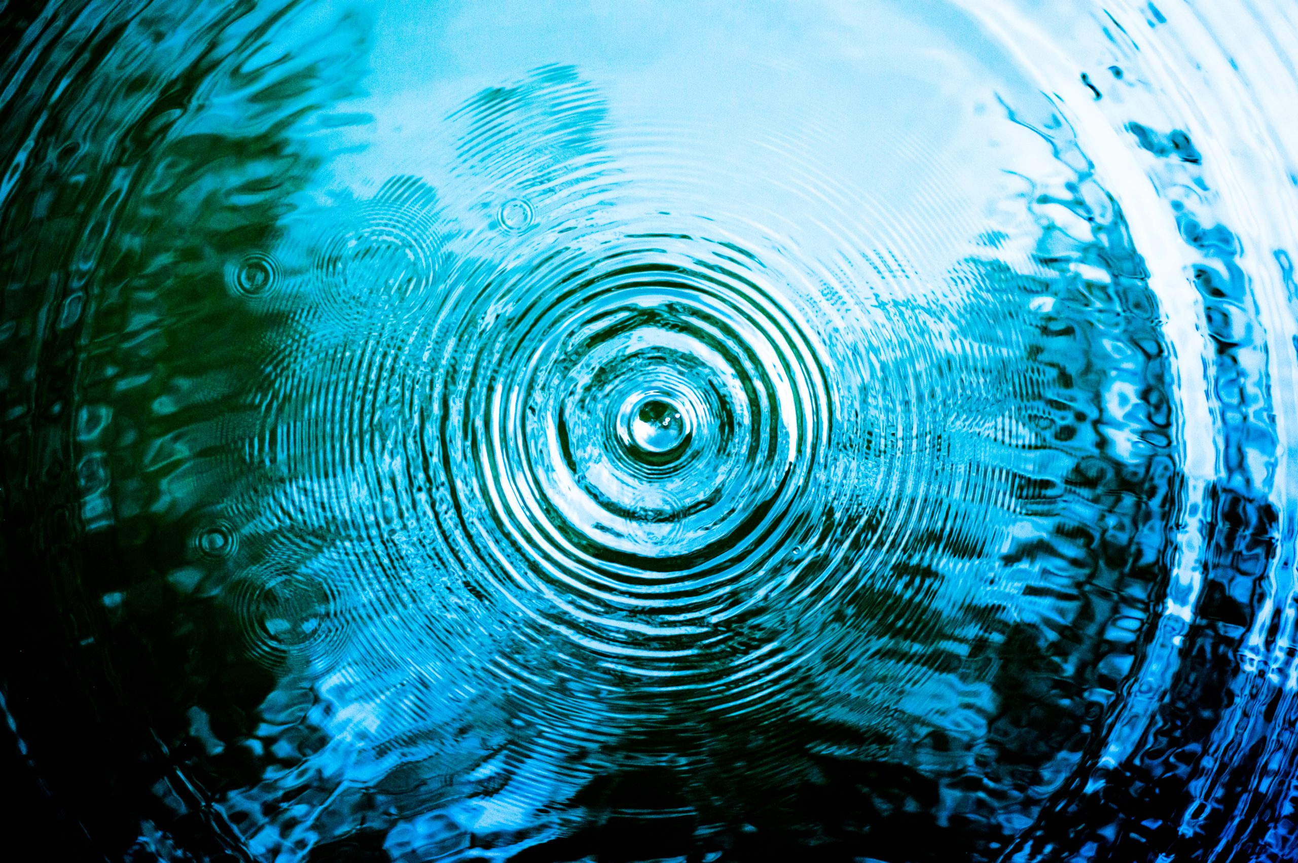 Concentric ripples spread across a tranquil blue water surface, reflecting light and subtle shadows.