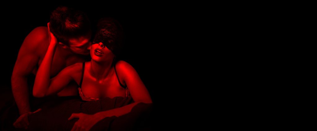 Couple in intimate pose, bathed in red light, with one partner blindfolded and the other leaning in closely.