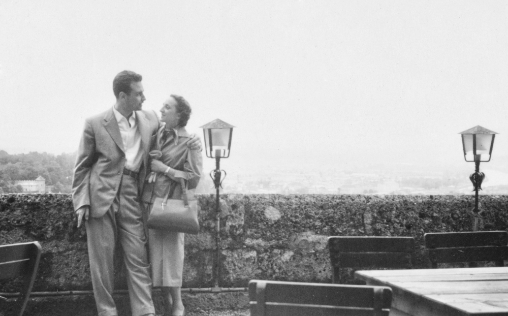 Couple in vintage attire gazes at each other, standing by a stone wall with lamps, overlooking a scenic view.