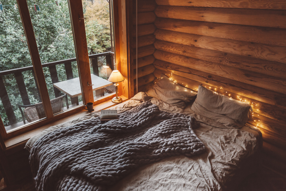 Cozy log cabin bedroom with a large bed, soft blanket, fairy lights, and a view of a balcony surrounded by trees.