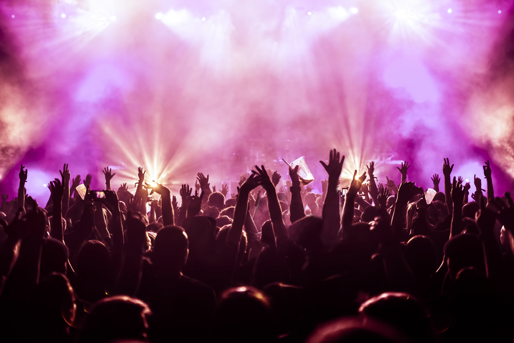 Crowd at a concert with raised hands, illuminated by vibrant purple and white lights, creating a lively atmosphere.