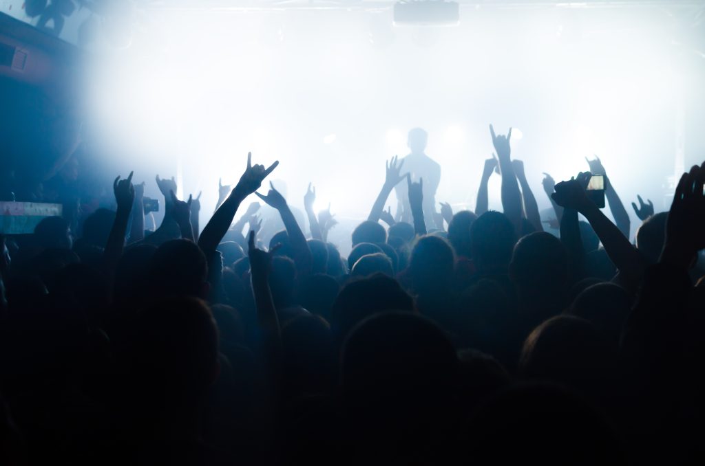 Crowd at a concert with raised hands, illuminated by bright stage lights and a hazy atmosphere.