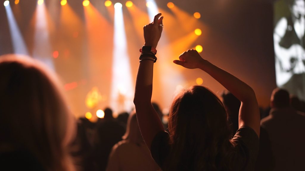 A crowd enjoying a concert, with a person raising their arms in excitement against vibrant stage lights.