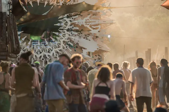 Crowd enjoying a music festival, with a DJ performing on a decorated stage surrounded by dust and vibrant energy.