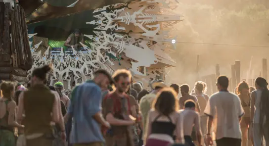 Crowd enjoying a music festival, with a DJ performing on a decorated stage surrounded by dust and vibrant energy.