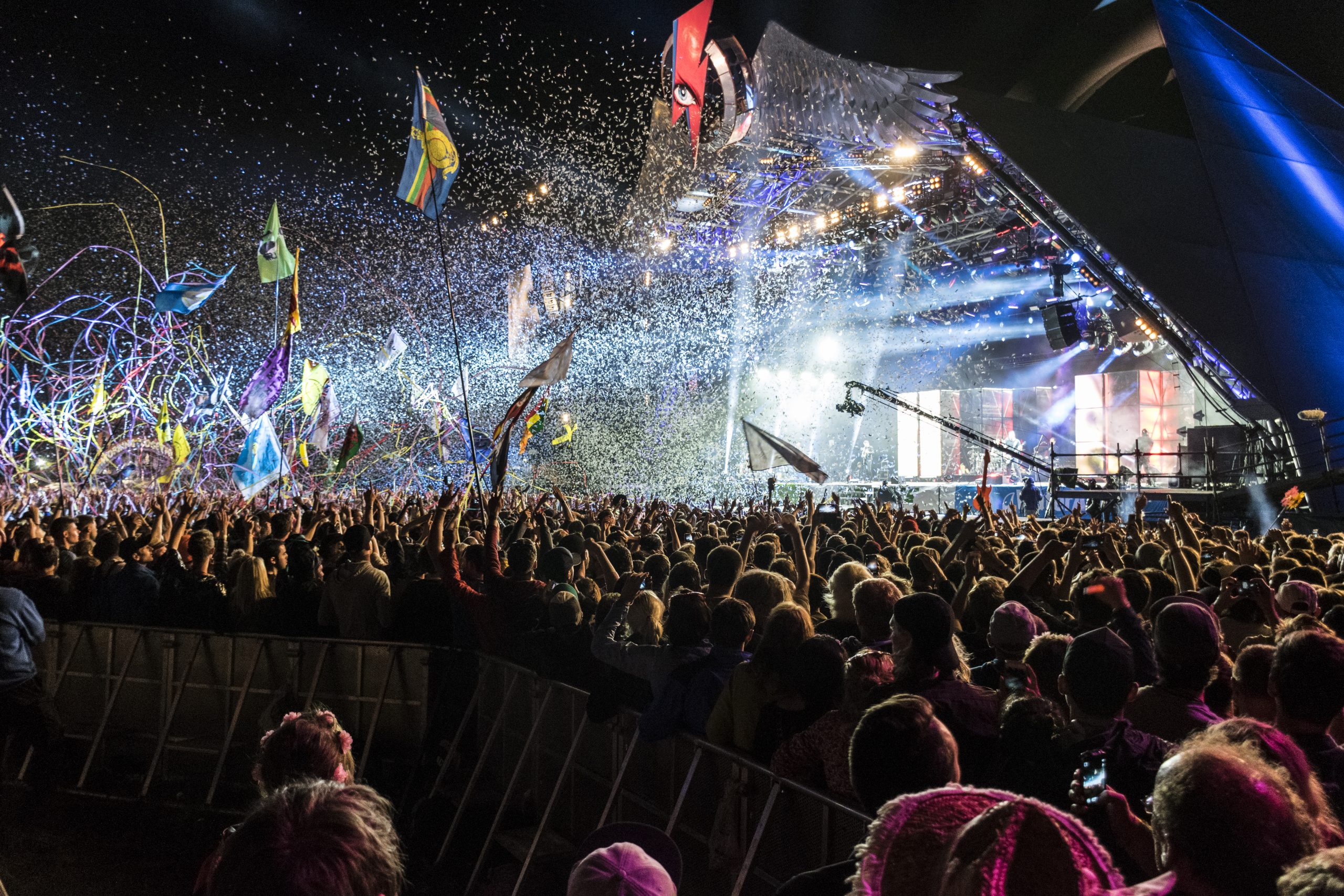 Crowd at a lively concert, celebrating with colorful confetti and flags under bright stage lights.