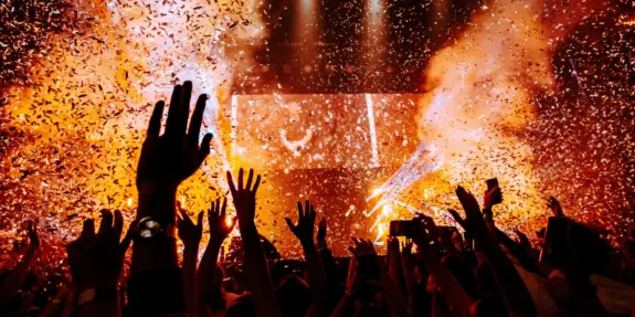 Crowd with raised hands celebrating amidst a shower of confetti and vibrant lights at a lively concert.