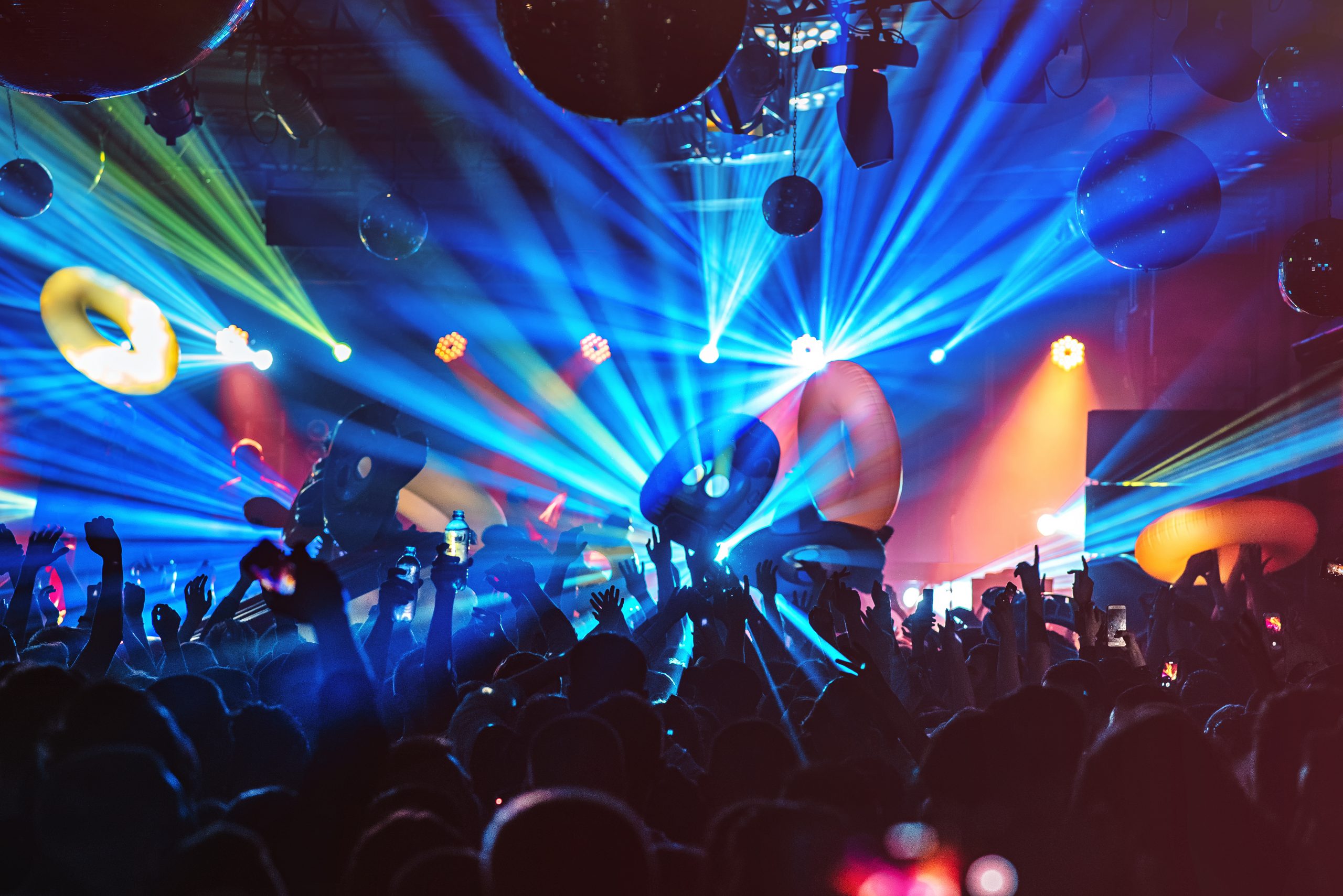 Crowd at a vibrant party, waving colorful inflatable rings under dynamic laser lights and disco balls.