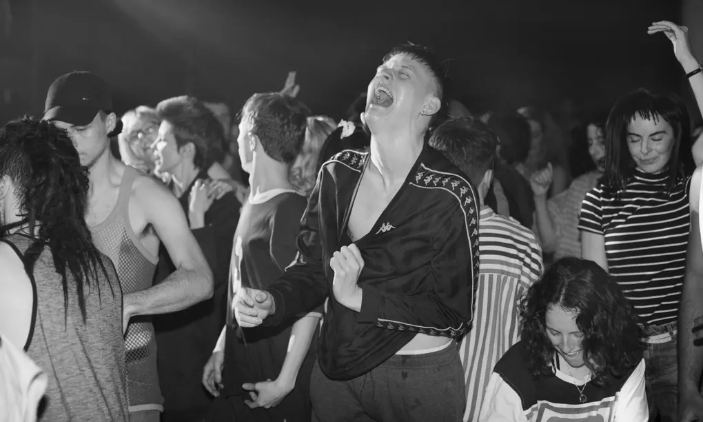 Crowd of young people dancing energetically at a concert, with expressions of joy and excitement. Black and white tones.