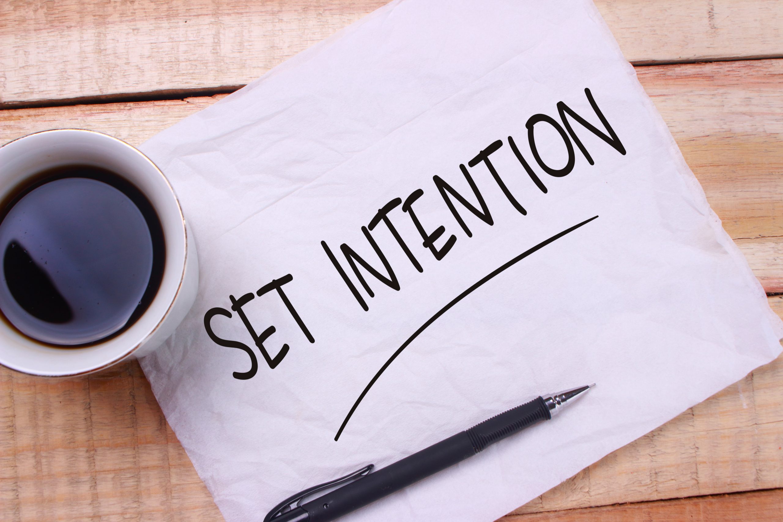 A cup of coffee beside a note that reads "SET INTENTION" on crumpled paper, resting on a wooden surface.