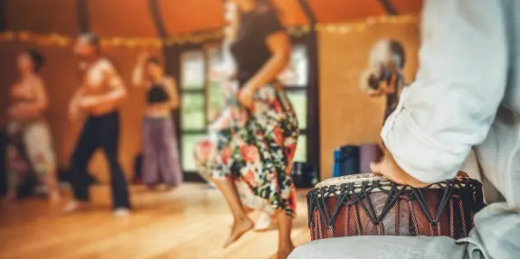 A dancer in a floral skirt moves energetically while a drummer plays in a lively, warm-toned studio.