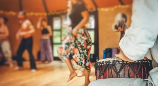 A dancer in a floral skirt moves energetically while a drummer plays in a lively, warm-toned studio.