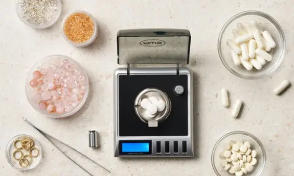 Digital scale with white capsules, surrounded by bowls of beads, crystals, and measuring tools on a light countertop.